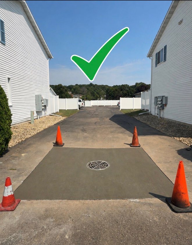 A full concrete pad poured around a storm grate set flush to grade in a driveway, coned off, marked with a green check