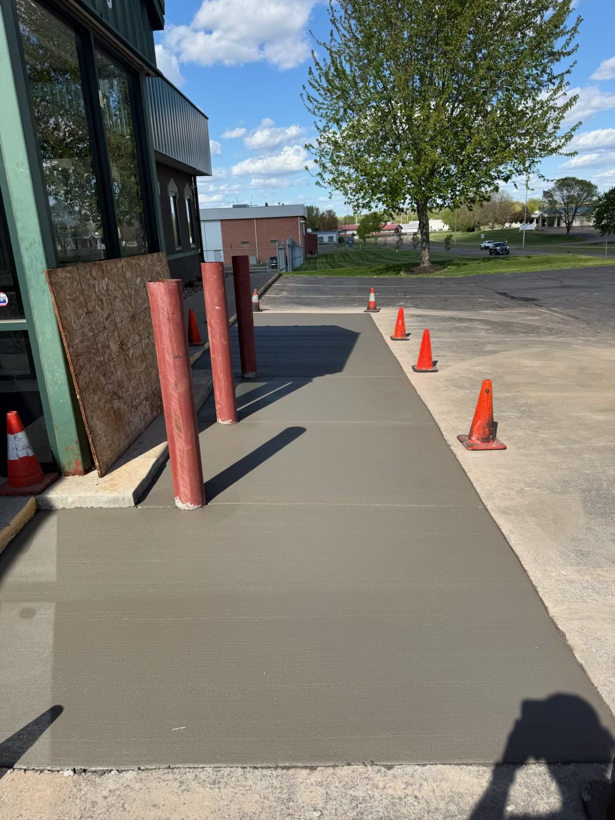 A freshly poured concrete walk running past steel bollards at a commercial storefront, coned off