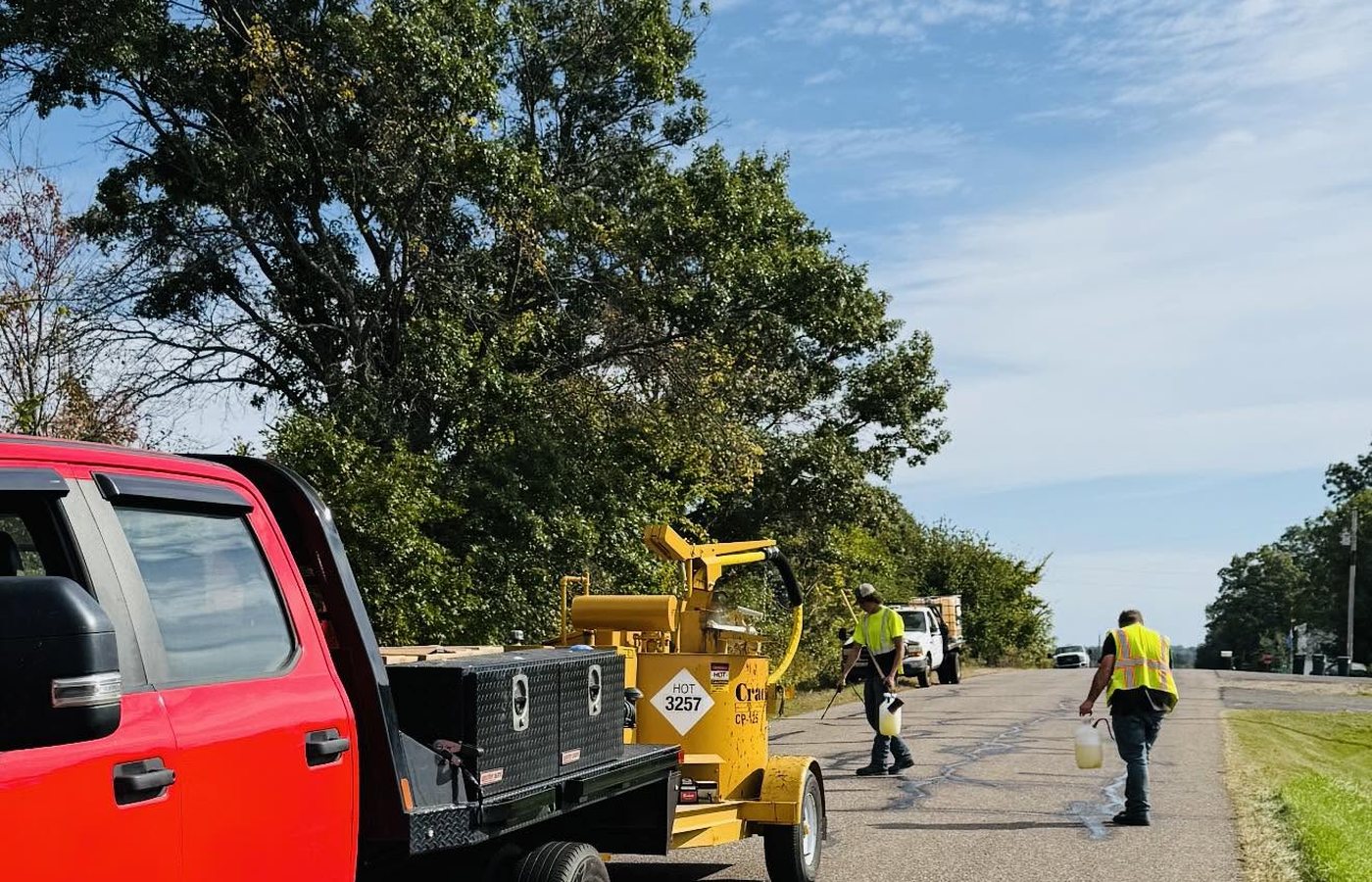The company flatbed and the yellow crack-seal melter on a road shoulder, two crew in hi-vis working ahead of it