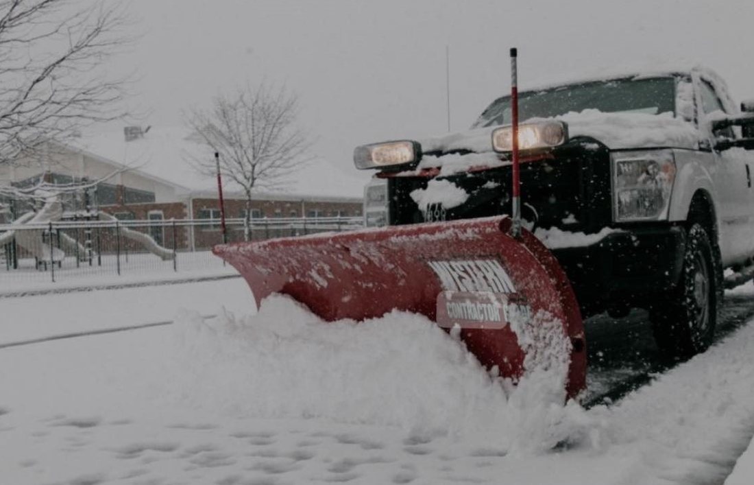 A plow truck with a red blade pushing a wave of snow across a commercial lot in daylight
